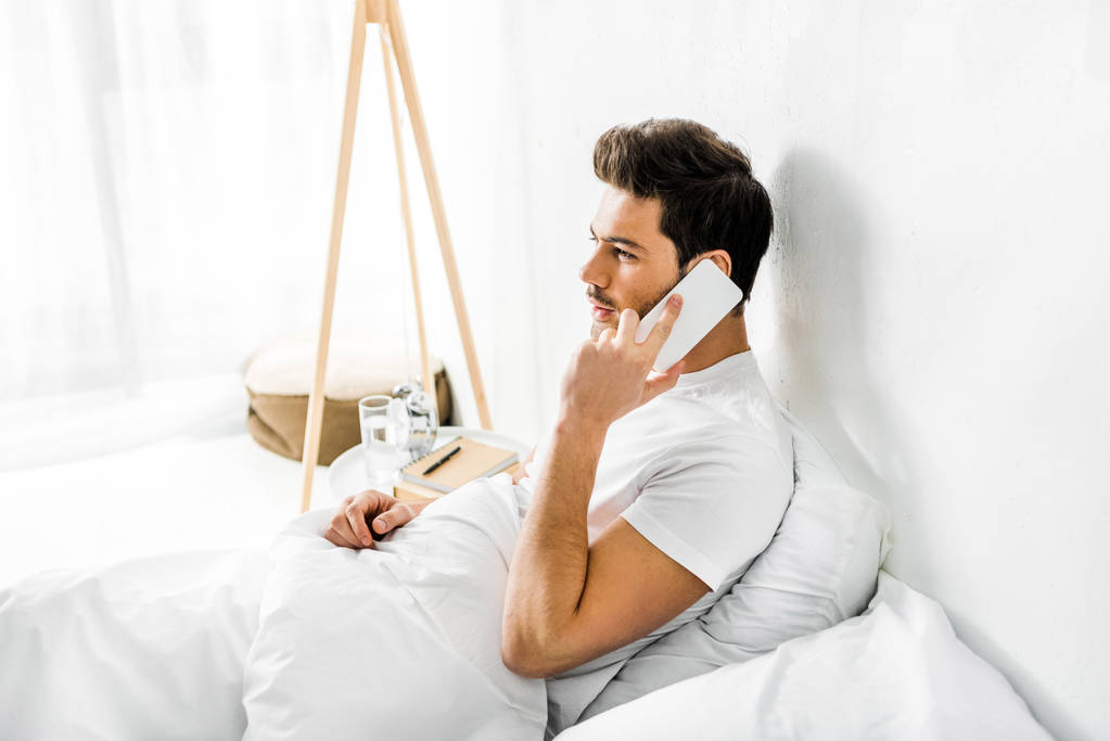 Nuori mies puhuu älypuhelimella istuessaan sängyssä aamulla
 - Valokuva, kuva