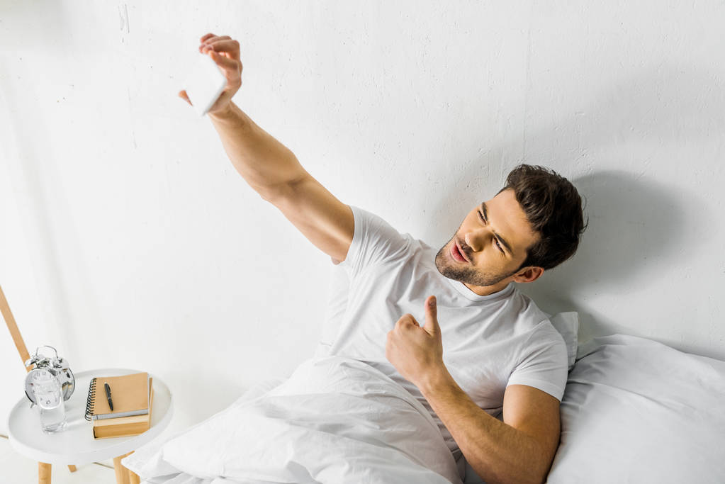 寝室でのスマート フォンの白を selfie を親指を示す幸せな男 - 写真・画像