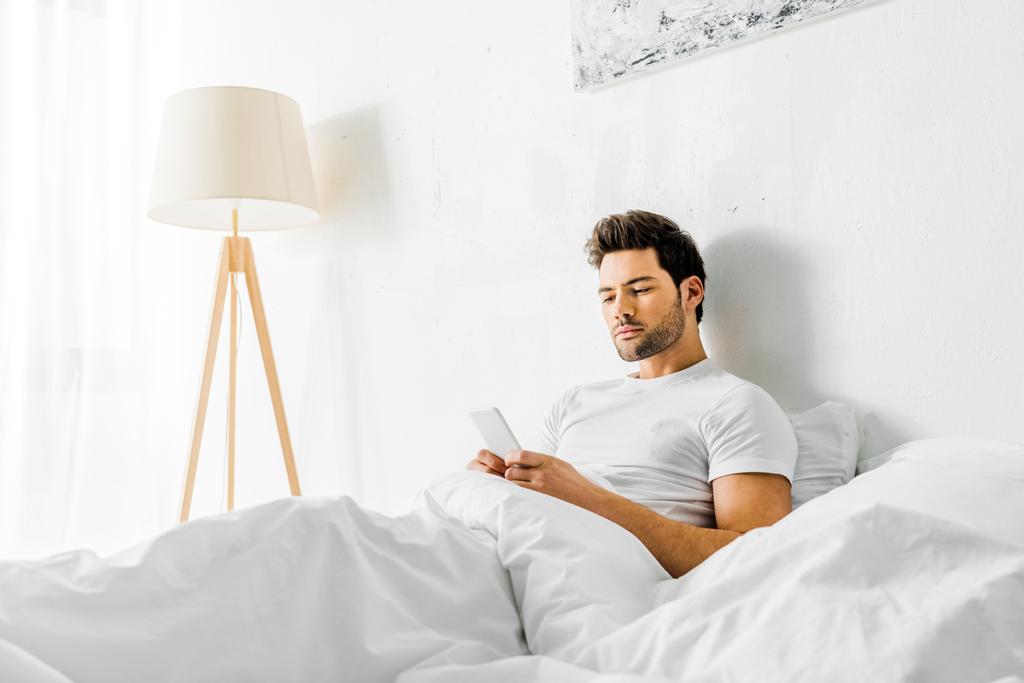 guapo hombre serio usando teléfono inteligente mientras descansa en la cama por la mañana
 - Foto, imagen