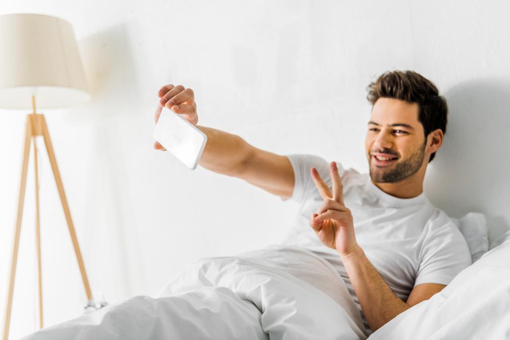 Lächelnder Mann zeigt Friedenssymbol beim Selfie mit Smartphone im Schlafzimmer - Foto, Bild