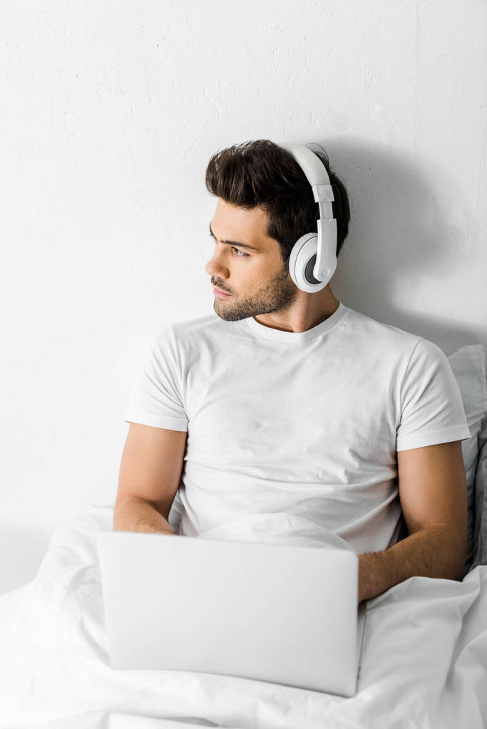 knappe man van de peinzende luisteren muziek met hoofdtelefoons en laptop in bed - Foto, afbeelding