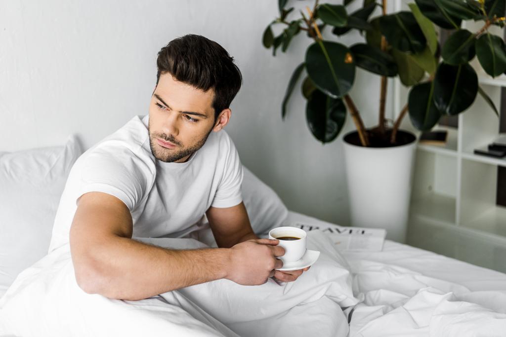 pensativo joven en pijama sosteniendo la taza de café en la cama
 - Foto, Imagen