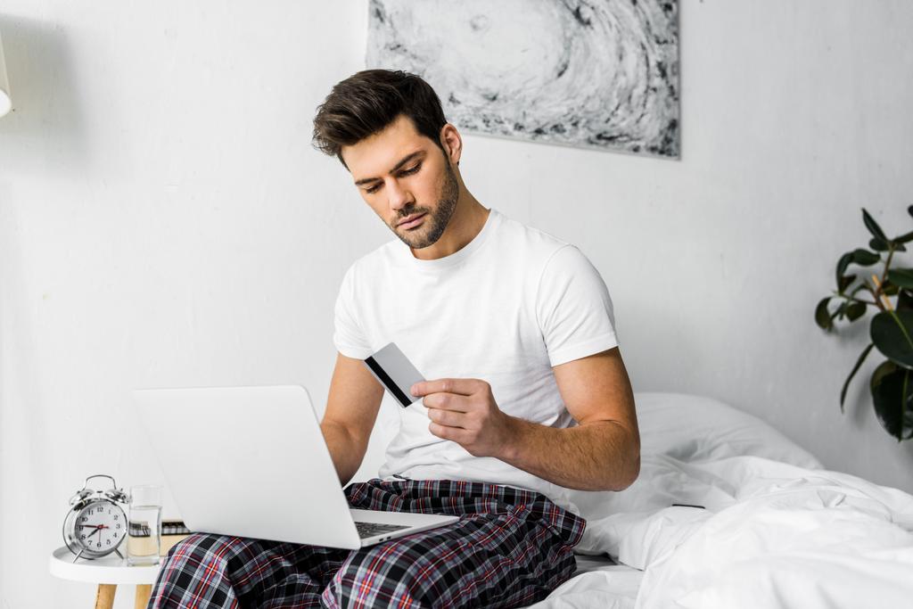 schöner Mann beim Online-Shopping mit Kreditkarte und Laptop im Schlafzimmer - Foto, Bild