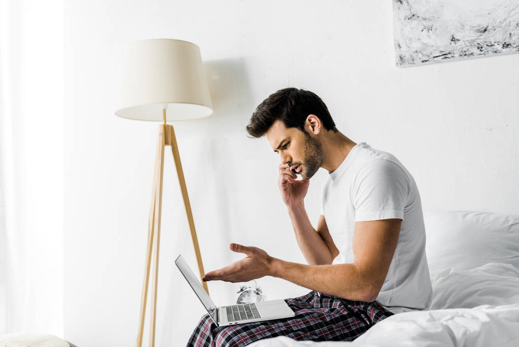 тривожний чоловік розмовляє на смартфоні і використовує ноутбук на ліжку
 - Фото, зображення