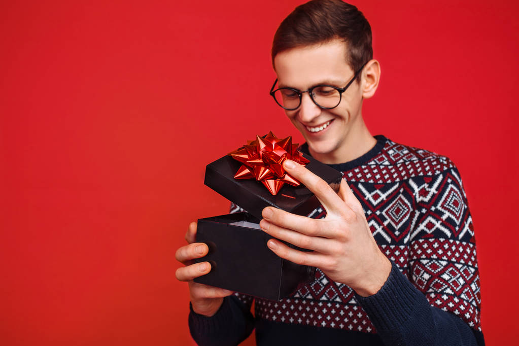 Чоловік в окулярах, який отримав подарунок, дивиться всередину коробки, на червоний фон
 - Фото, зображення