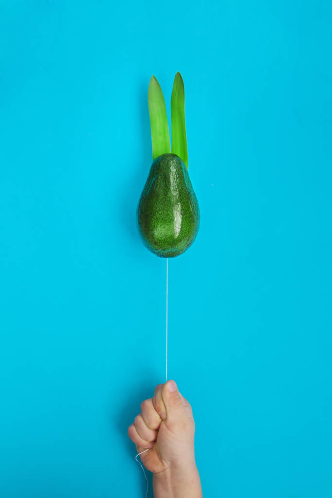 Beautiful creative decoration background idea with child's hand holding ropes flying balloon green avocado rabbit on bright blue paper. Vibrant spring card concept. Close up.Front view. Copy space. - Photo, Image