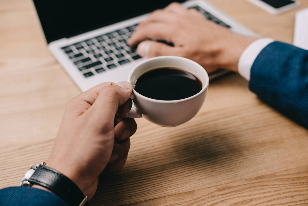 Teilansicht eines Geschäftsmannes, der am Arbeitsplatz eine Tasse Kaffee hält, während er am Laptop tippt - Foto, Bild