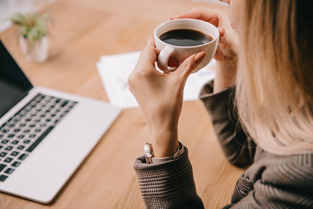 bijgesneden weergave van zakenvrouw houden van kopje koffie op kantoor met laptop - Foto, afbeelding