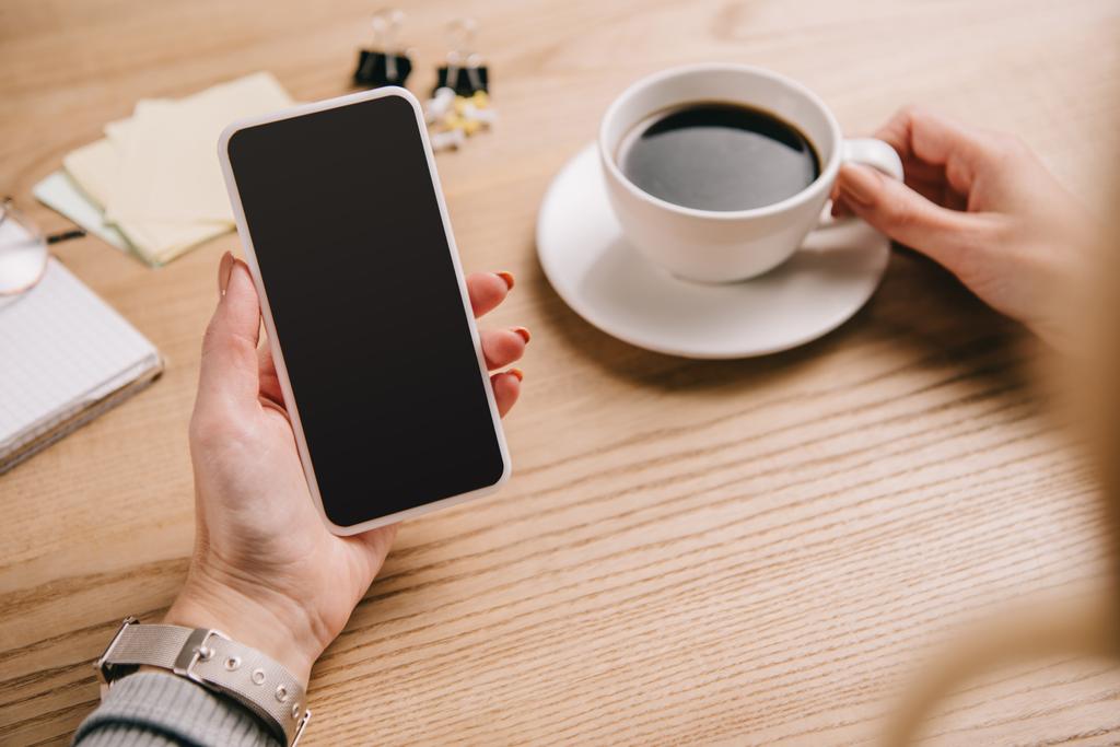 abgeschnittene Ansicht einer Geschäftsfrau, die eine Tasse Kaffee hält, während sie ihr Smartphone am Arbeitsplatz benutzt - Foto, Bild