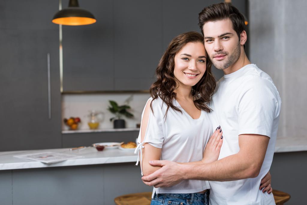 feliz pareja mirando a la cámara y abrazándose en la cocina
  - Foto, Imagen