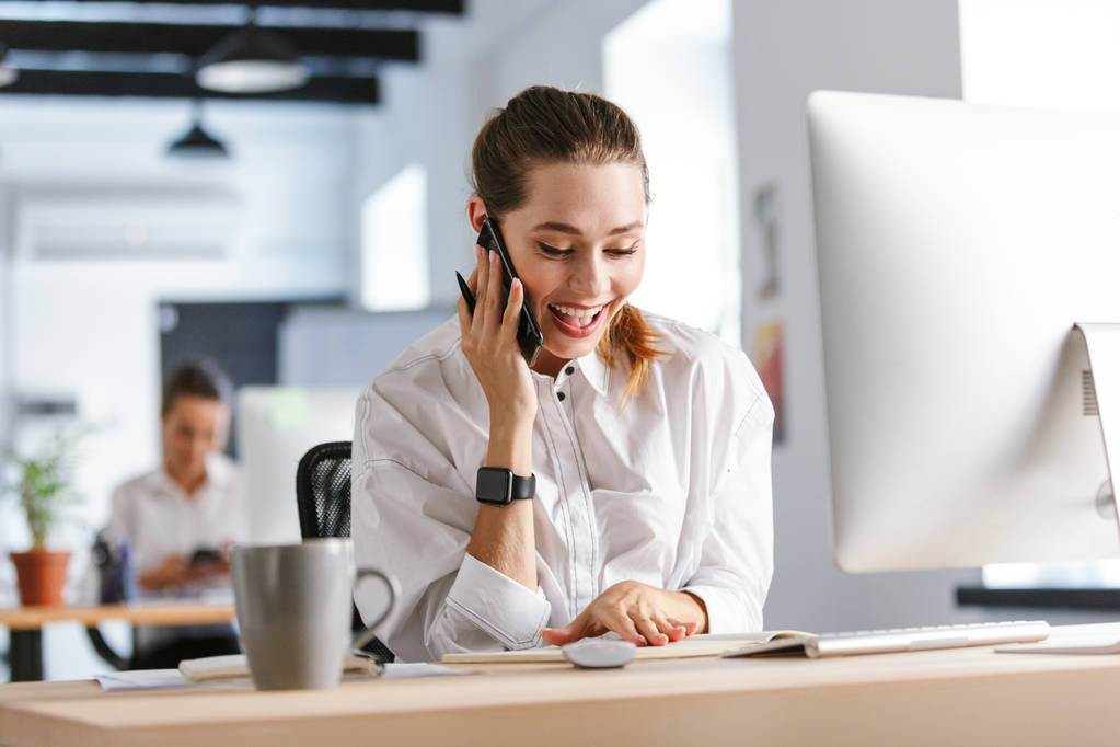 Χαρούμενος νεαρός επιχειρηματίας κάθεται στο χώρο εργασίας της στο γραφείο, μιλώντας σε κινητό τηλέφωνο, λήψη σημειώσεων - Φωτογραφία, εικόνα