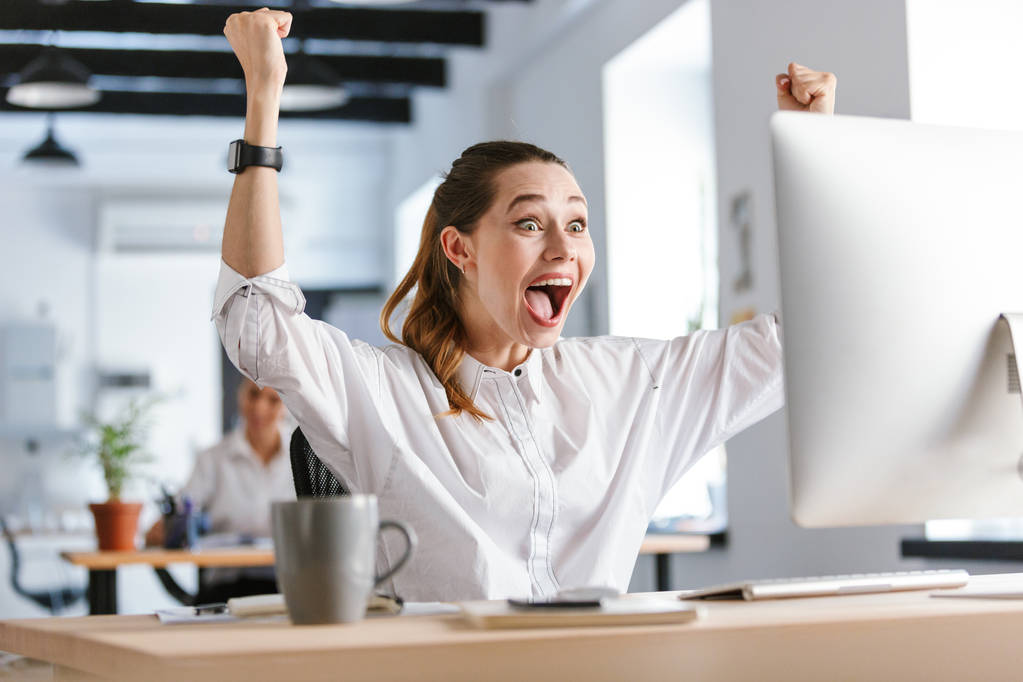 Χαρούμενος νεαρός επιχειρηματίας κάθεται στο χώρο εργασίας της στο γραφείο, να γιορτάζει την επιτυχία, να βλέπουν τον υπολογιστή - Φωτογραφία, εικόνα