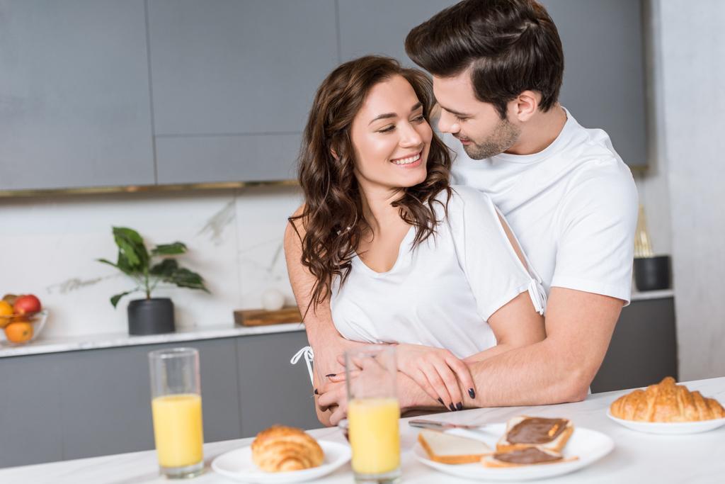ευτυχισμένο ζευγάρι αγκαλιάζει και χαμογελαστός στην κουζίνα κοντά νόστιμο πρωινό - Φωτογραφία, εικόνα