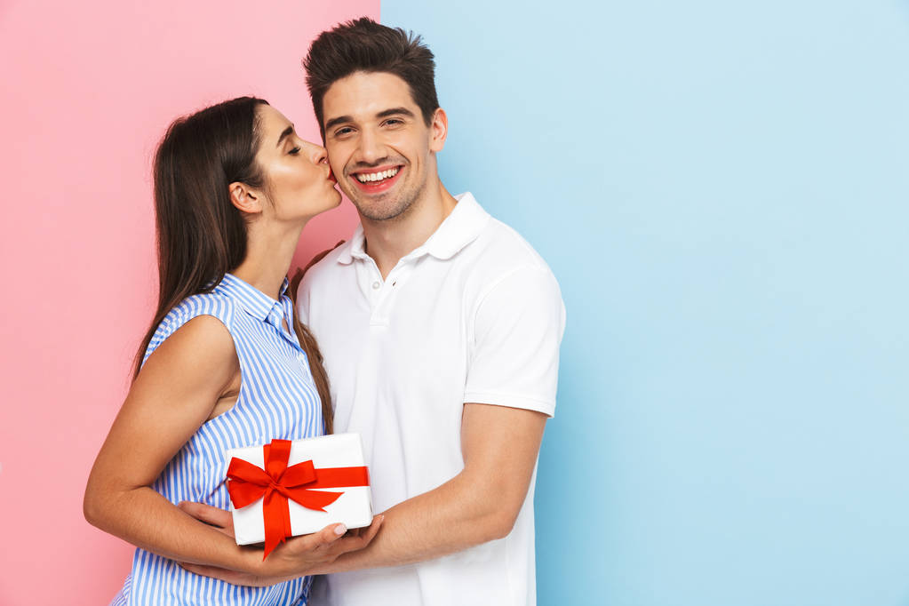 glückliches junges Paar steht isoliert auf zwei farbigen Hintergrund, feiert, hält Geschenkbox, küsst - Foto, Bild