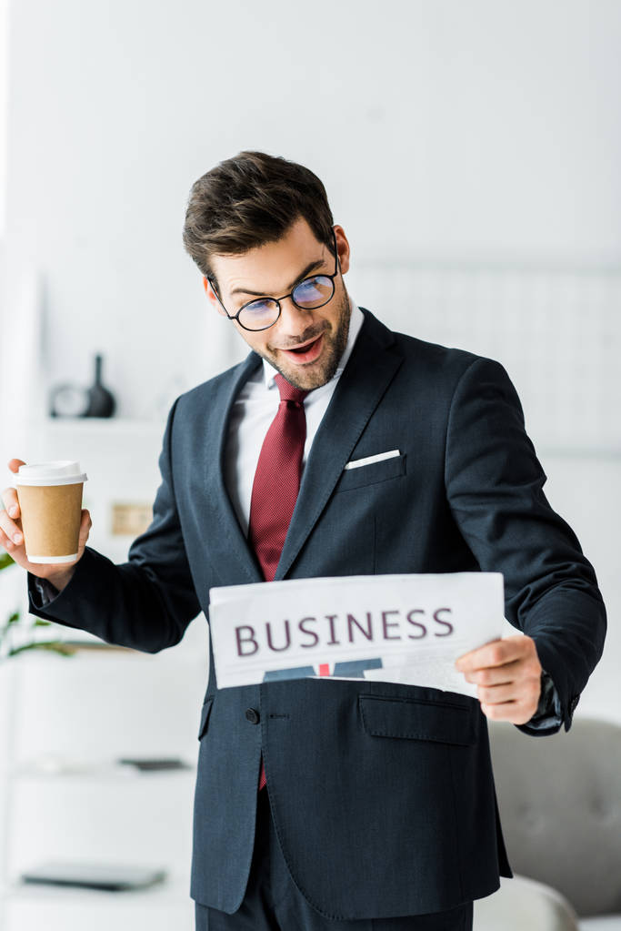 surprised businessman in formal wear holding coffee to go and reading newspaper in office - Foto, afbeelding