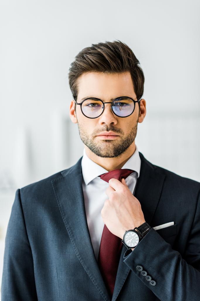 handsome businessman in formal wear and glasses adjusting tie while looking at camera in office - Photo, Image