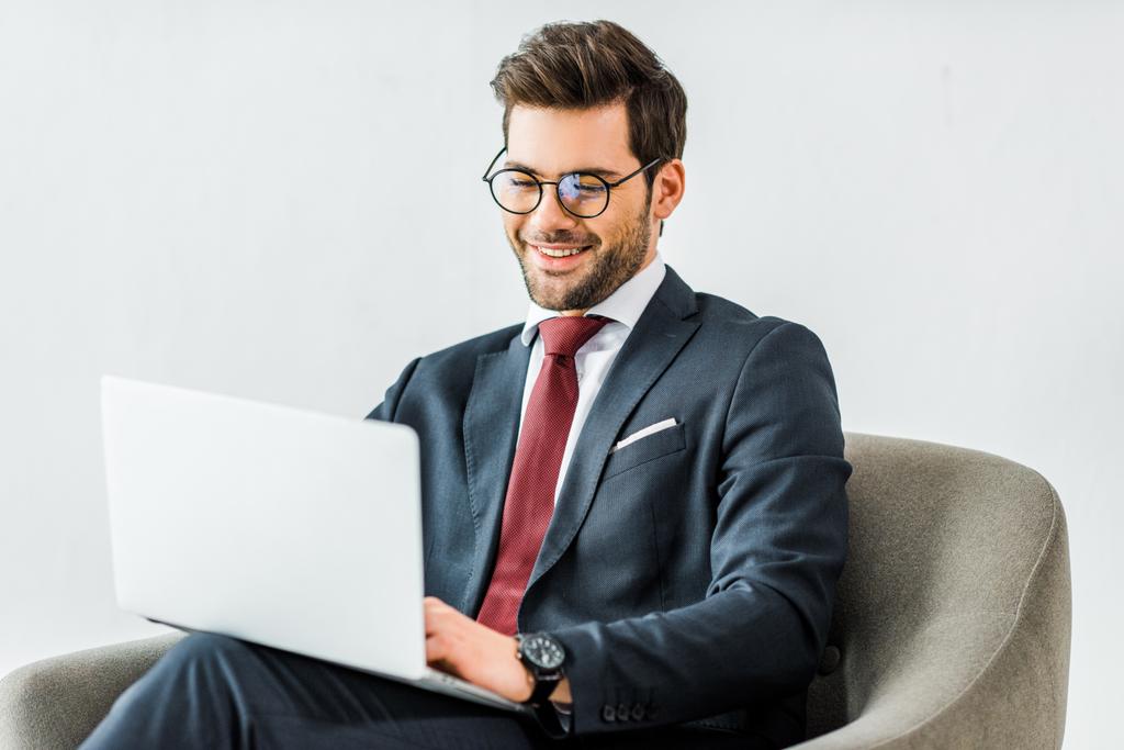 Lächelnder Geschäftsmann in offizieller Kleidung sitzt auf Sessel und benutzt Laptop im Büro - Foto, Bild