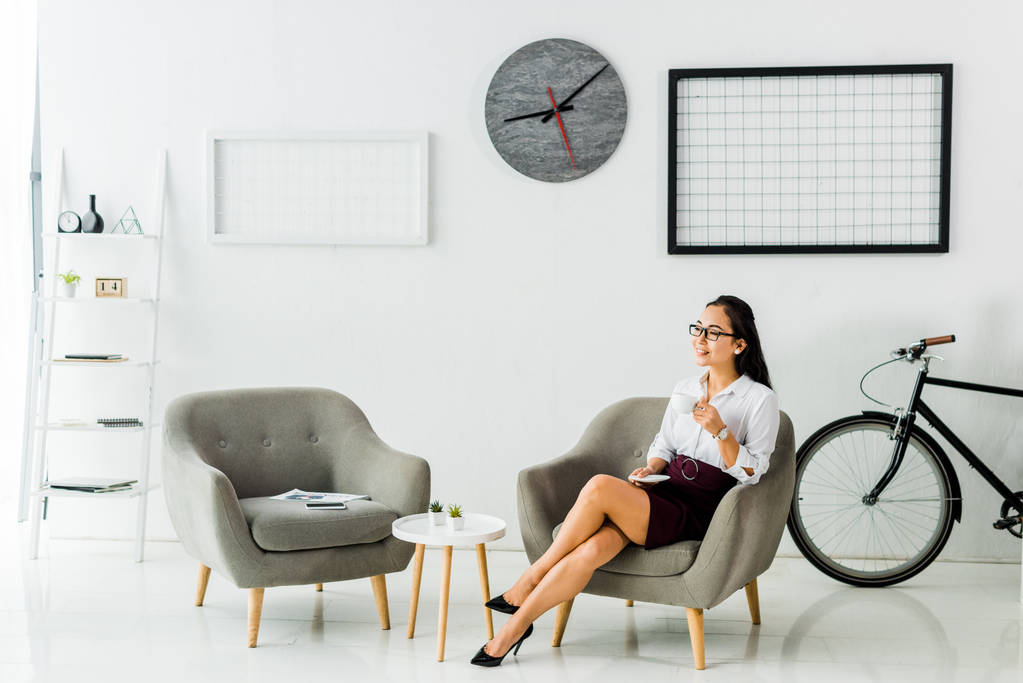 souriant asiatique femme d'affaires assis sur fauteuil avec tasse de café dans le bureau moderne
  - Photo, image