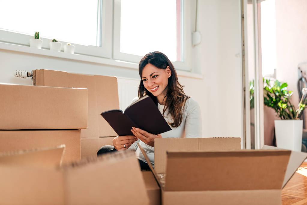 Attractive young woman is moving, reading book and smiling while packing. - Photo, Image