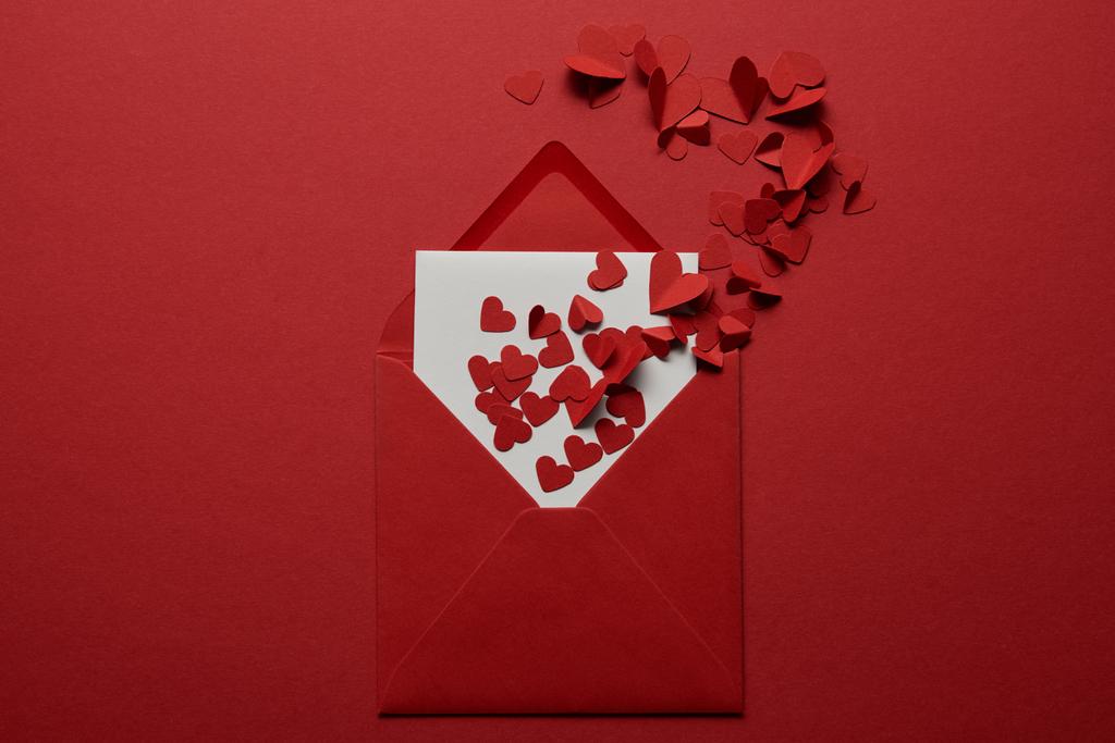 Draufsicht auf weißen Brief im Umschlag mit Stapel aus Papier geschnittene Herzen auf rotem Hintergrund - Foto, Bild