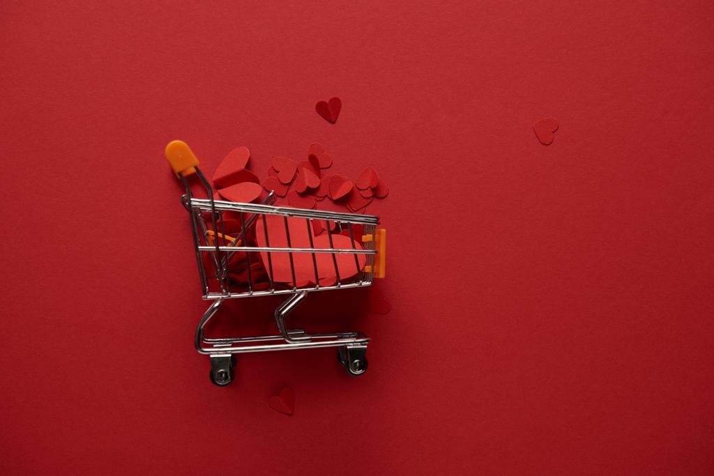 top view of toy shopping cart with decorative paper cut hearts on red background - Photo, Image