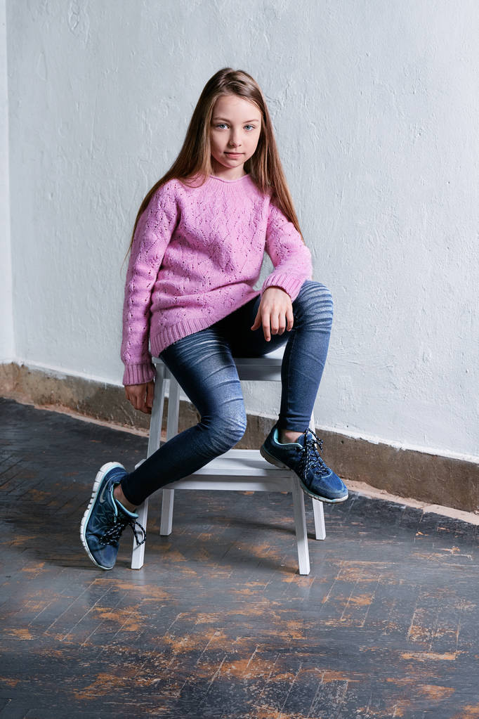 Itsevarma lapsi tyttö istuu muoti aiheuttaa tuolilla, betoni valkoinen tausta. Tyylikäs malli vaaleanpunainen pusero. Kaunis lumoava lapsi teini, rento, kaupunkien nuorten tyyli, vaatteet. Studiokuva
. - Valokuva, kuva