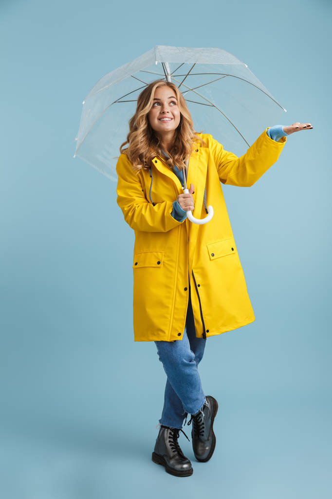 Ganzkörperbild einer blonden Frau in den 20er Jahren, die einen gelben Regenmantel trägt und unter einem transparenten Regenschirm steht, der über blauem Hintergrund isoliert ist - Foto, Bild