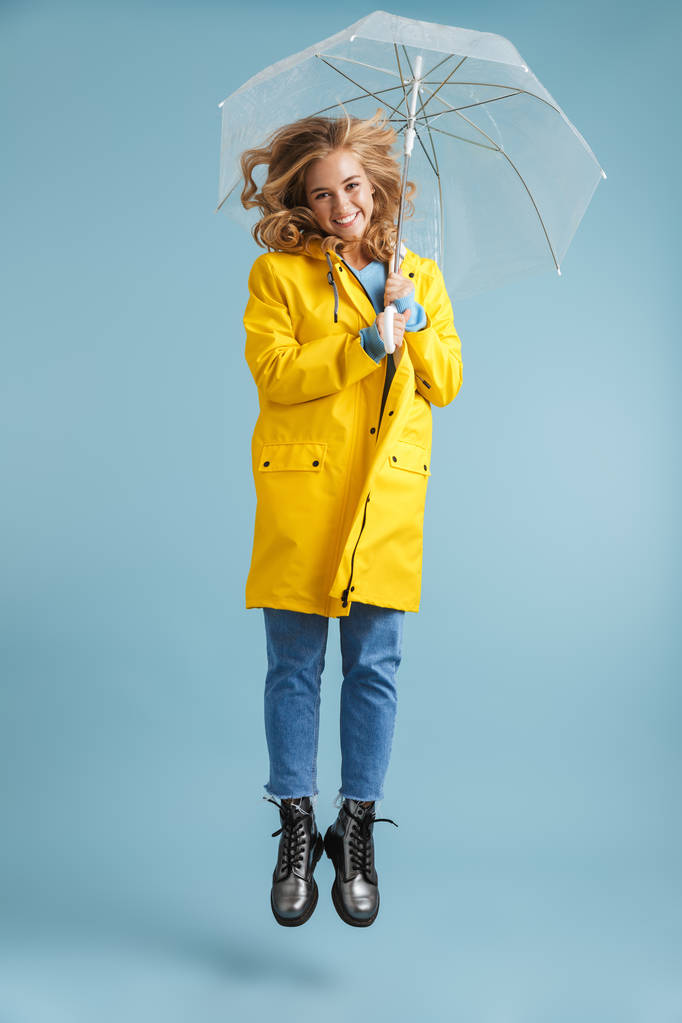 Ganzkörperbild einer positiven Frau in den 20er Jahren, die einen gelben Regenmantel trägt und unter einem transparenten Regenschirm steht, der über blauem Hintergrund isoliert ist - Foto, Bild