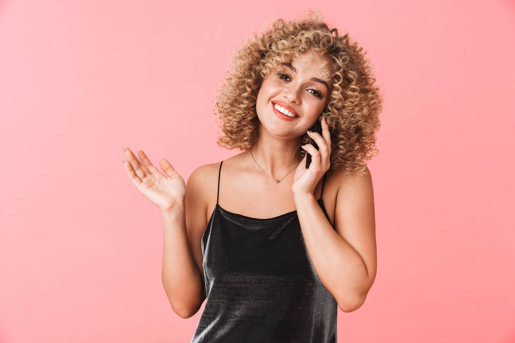 Portrét okouzlující kudrnaté ženy 20s usmívající se a mluvit na mobilním telefonu, samostatný růžové pozadí - Fotografie, Obrázek