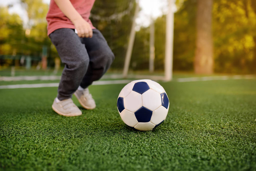 Мальчик веселится, играя в футбол / футбол в летний день. Активная игра на открытом воздухе / спорт для детей. Детские футбольные курсы и лагеря
 - Фото, изображение