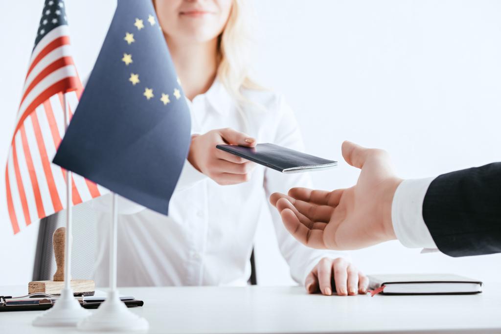cropped view of woman giving passport to tourist near american and european flags isolated on white - Photo, Image