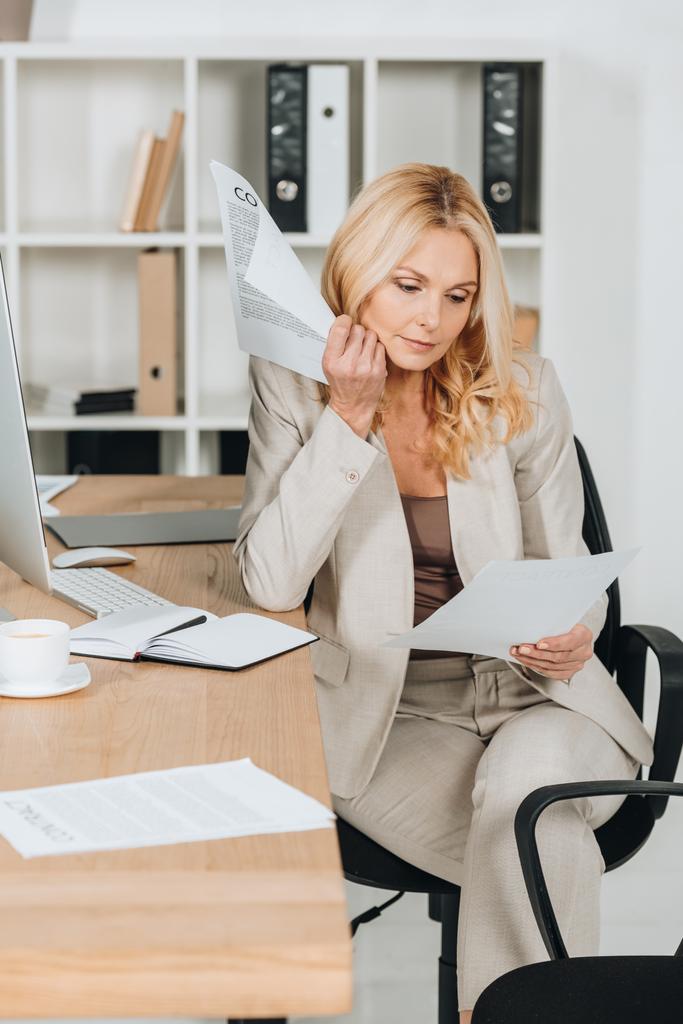 серйозна зріла бізнес-леді, що працює з паперами, сидячи на робочому місці
 - Фото, зображення