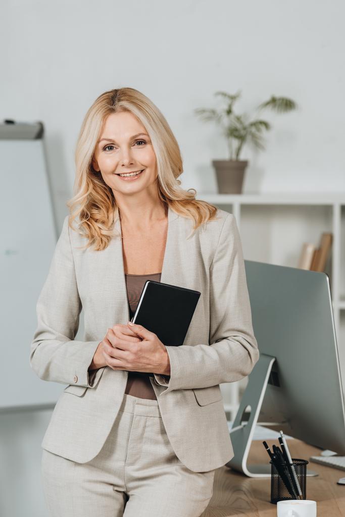 beautiful mature businesswoman holding notebook and smiling at camera in office - Photo, Image