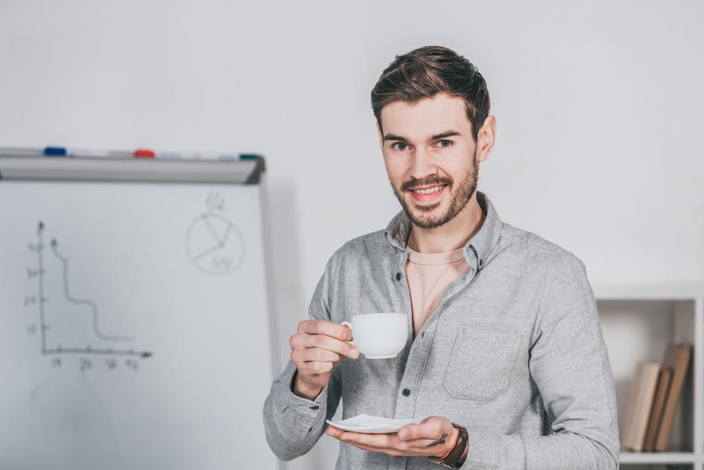 guapo joven empresario sosteniendo la taza de café y sonriendo a la cámara en la oficina
  - Foto, Imagen