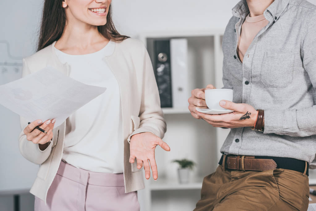 обрізаний знімок усміхненої бізнес-леді, що тримає папери та колегу-чоловіка, що тримає чашку кави в офісі
  - Фото, зображення