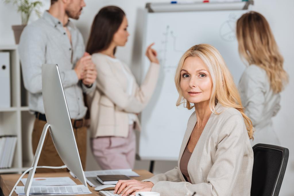 mature femme d'affaires en utilisant un ordinateur de bureau et en regardant la caméra tandis que les jeunes collègues travaillent avec tableau blanc derrière
 - Photo, image