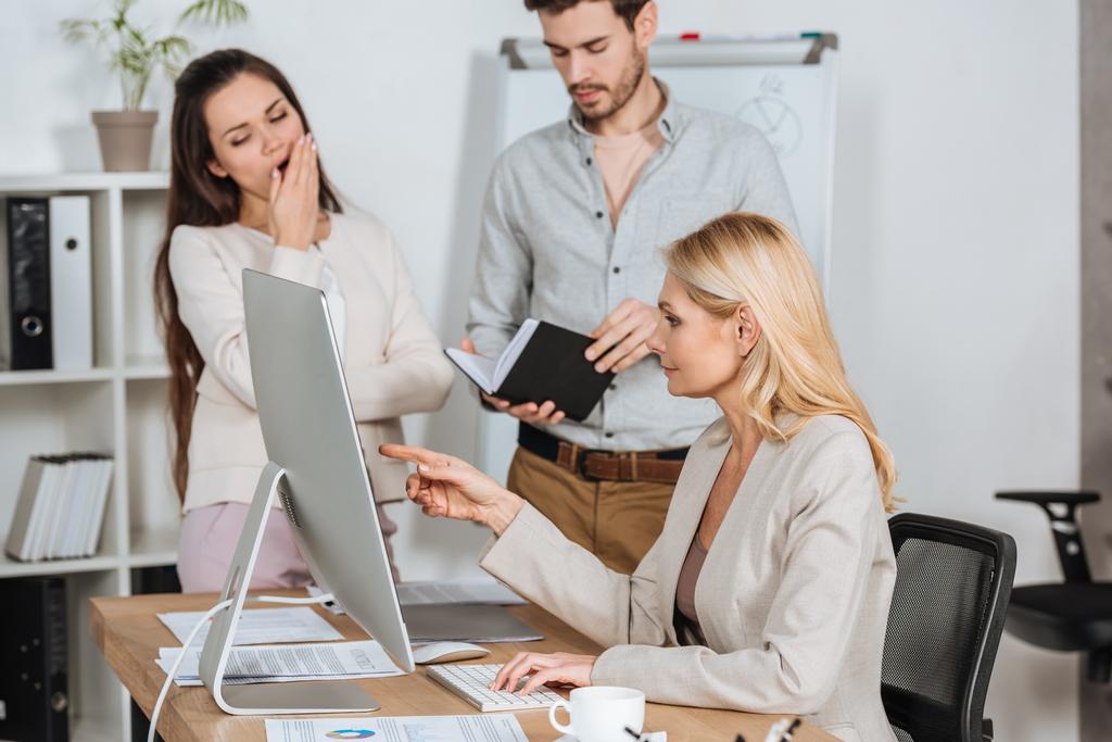 professionele mentor wijzend op de desktopcomputer en werken met verveeld jonge collega's in office - Foto, afbeelding