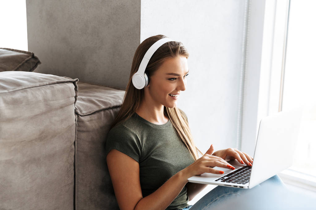 Εύθυμη νεαρή γυναίκα να ακούτε μουσική με ακουστικά στο σπίτι, λειτουργεί σε φορητό υπολογιστή - Φωτογραφία, εικόνα