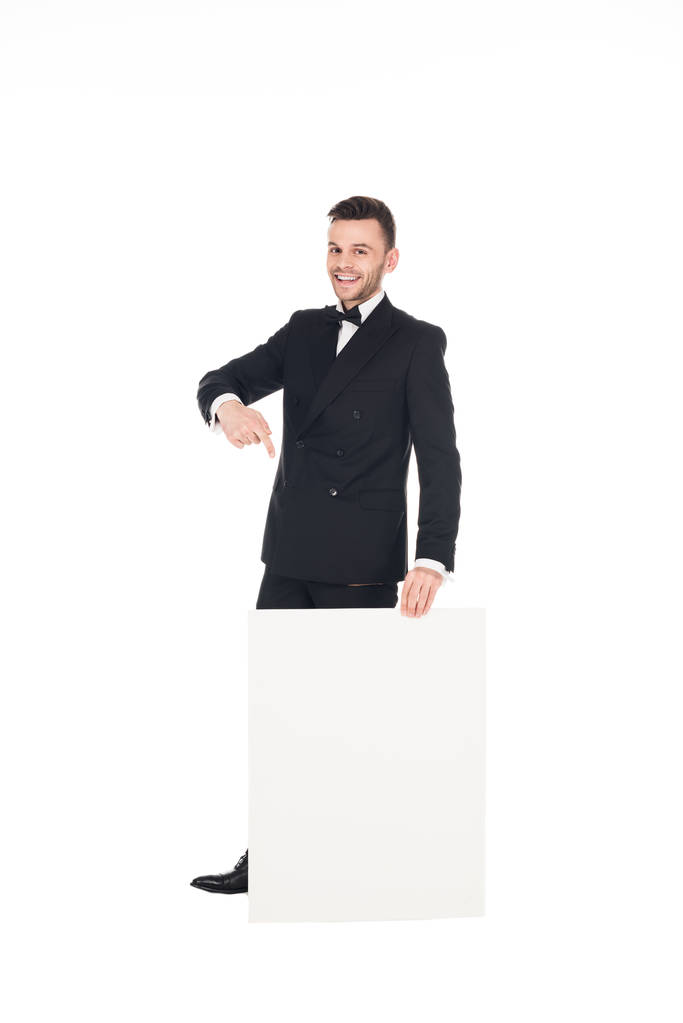 vrolijke elegante man in zwart pak wijzend op leeg bord geïsoleerd op wit - Foto, afbeelding