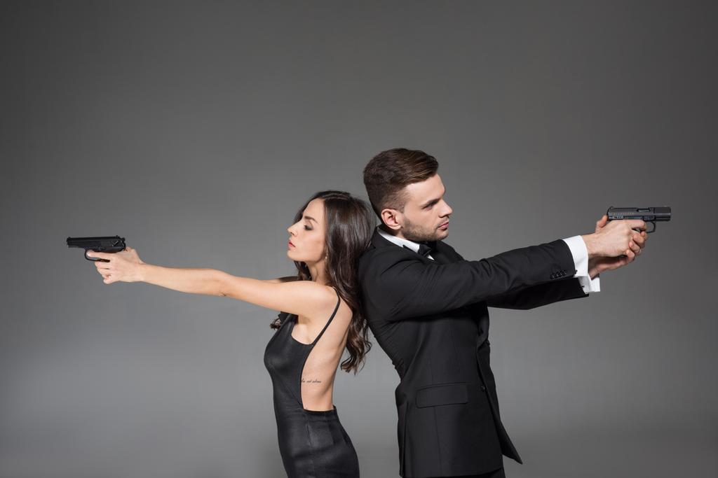 confiante casal de agentes secretos em roupas pretas visando com arma, isolado em cinza
 - Foto, Imagem
