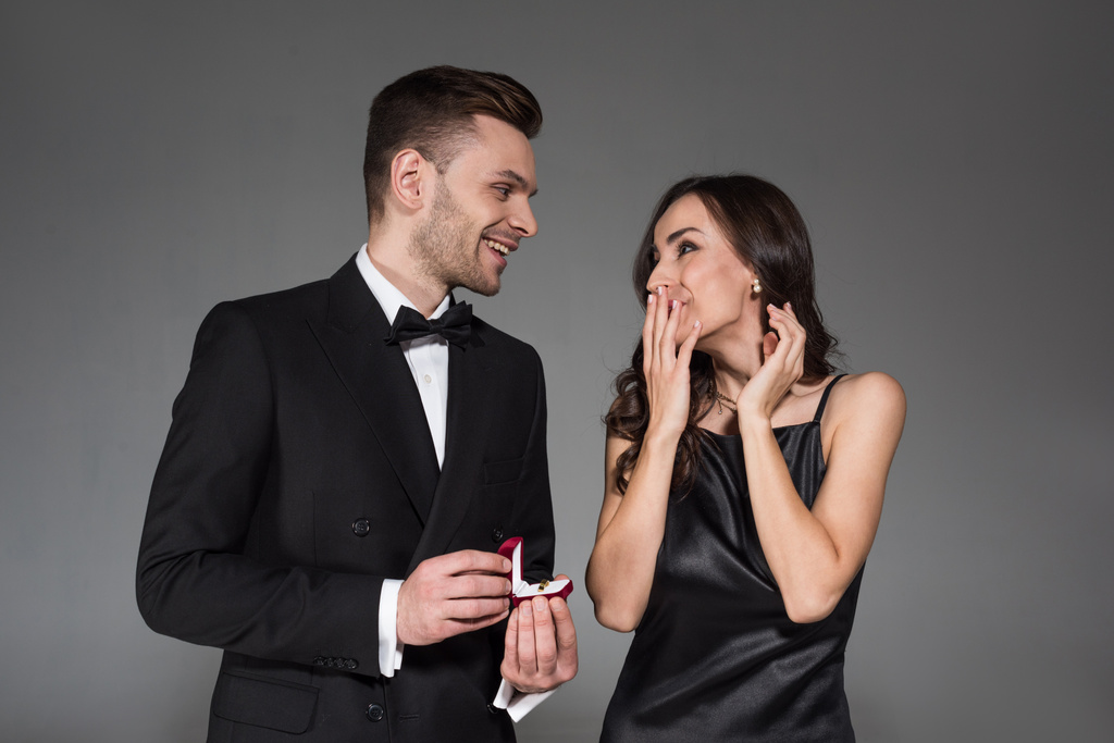 schöner Freund hält Heiratsantrag Ring in Box für aufgeregte Freundin, isoliert auf grau - Foto, Bild