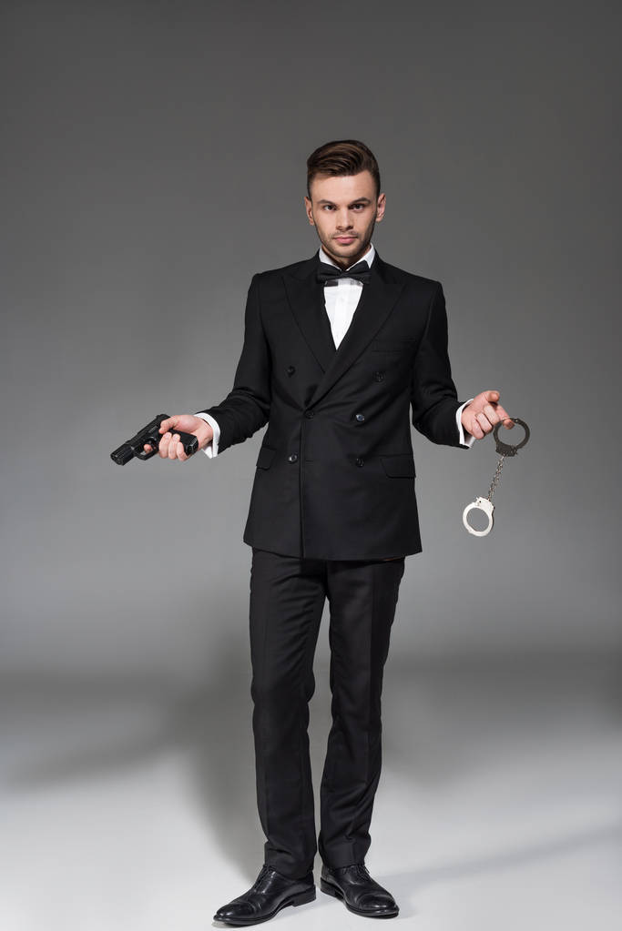 elegant secret agent in tuxedo holding handgun and handcuffs on grey - Photo, Image