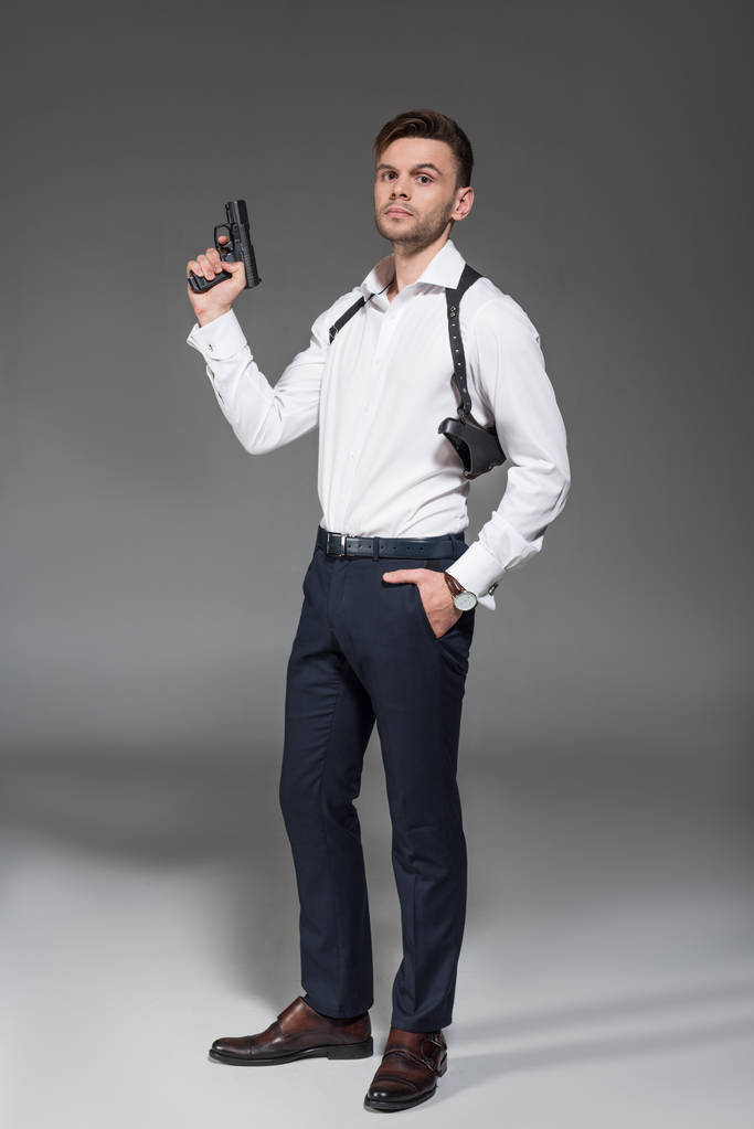 guapo joven agente secreto con funda sosteniendo pistola en gris
 - Foto, imagen
