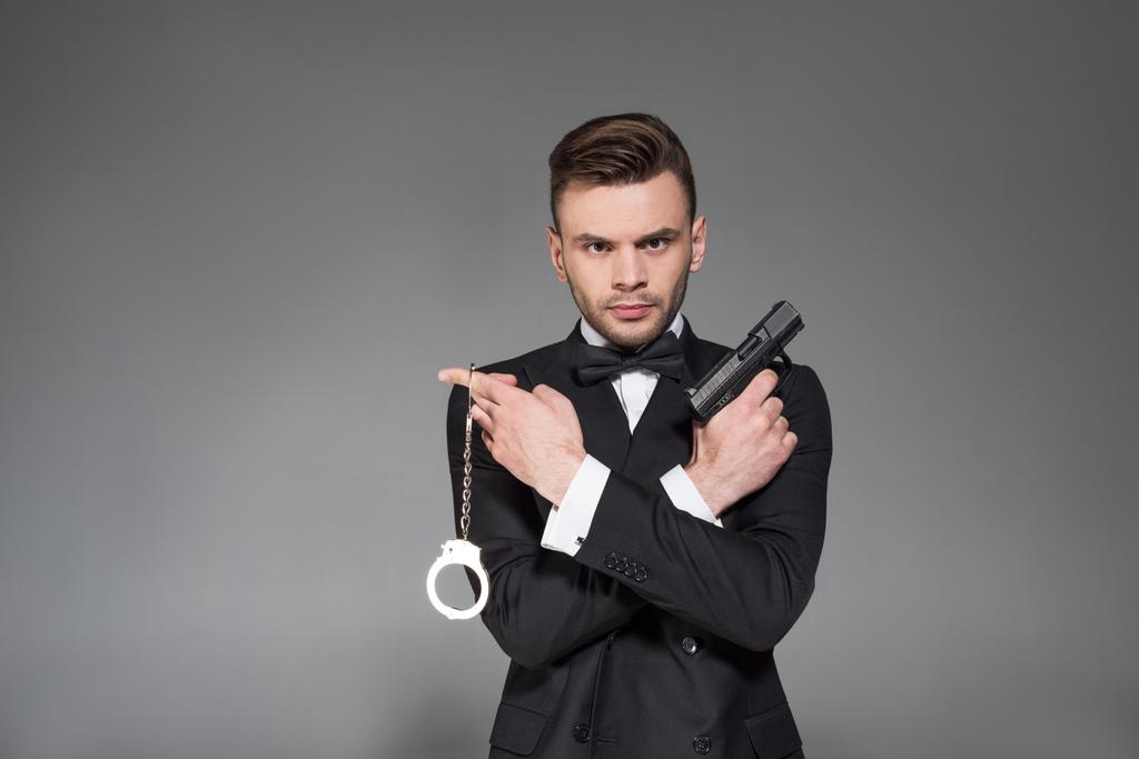 serious secret agent in black tuxedo holding gun and handcuffs, isolated on grey - Photo, Image