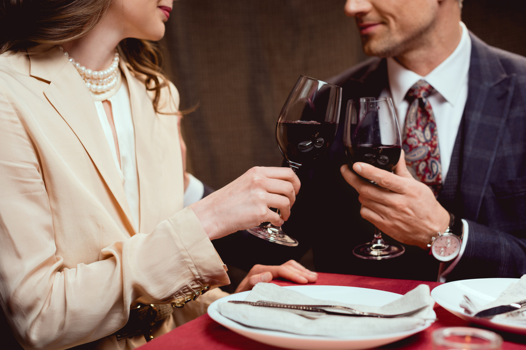 částečný pohled pár cinkání sklenic červeného vína během romantické rande v restauraci - Fotografie, Obrázek