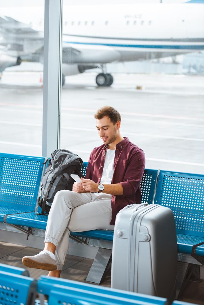 Χαμογελαστός άνθρωπος χρησιμοποιώντας το smartphone, ενώ κάθεται στην αίθουσα αναμονής στο αεροδρόμιο - Φωτογραφία, εικόνα