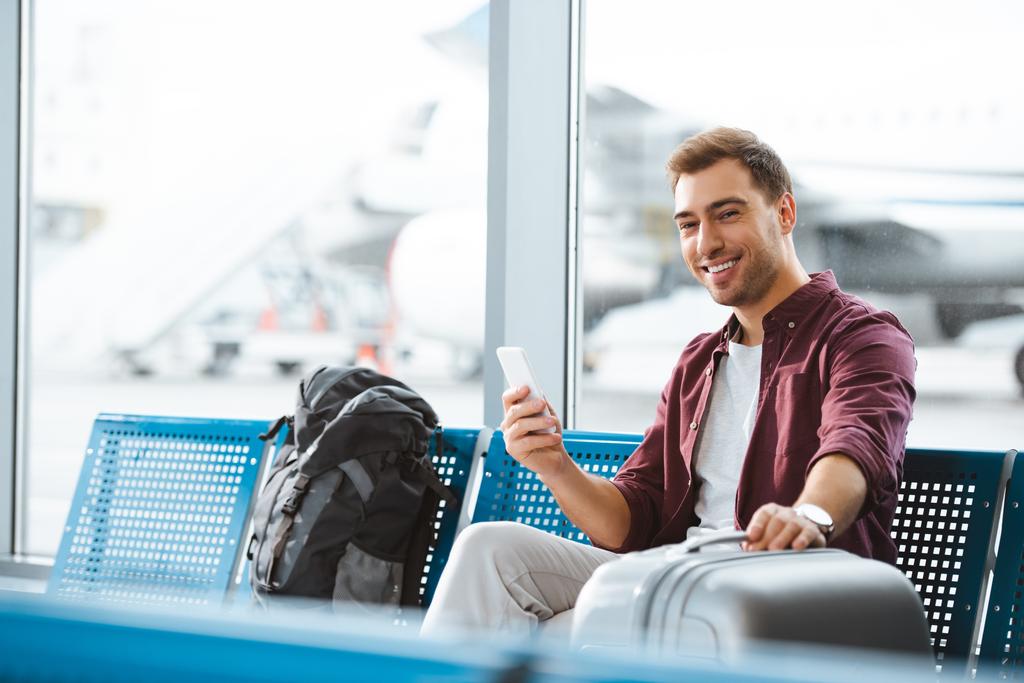 cheerful man looking at camera while holding smartphone and luggage in departure lounge - Photo, Image