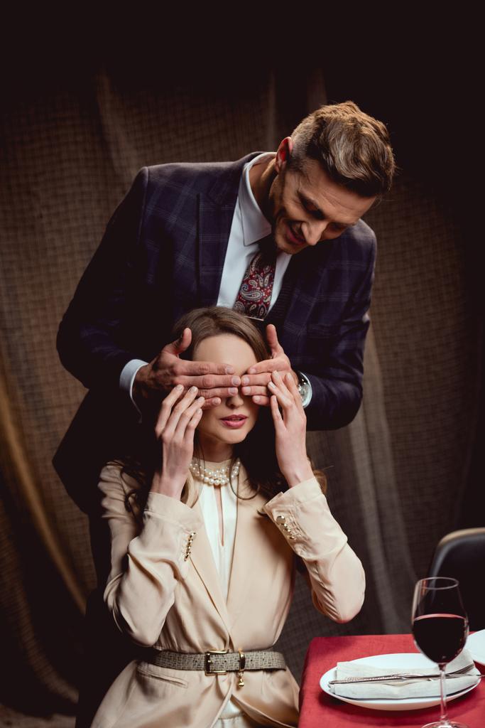 Улыбающийся мужчина закрывает глаза женщины руками для сюрприза во время романтического свидания в ресторане
 - Фото, изображение