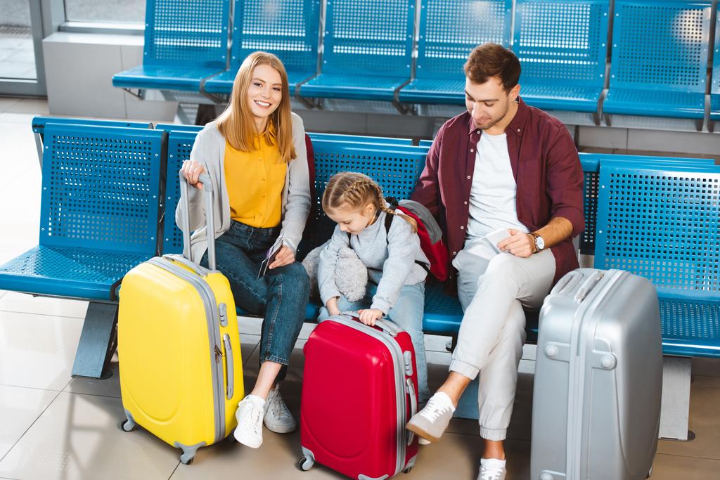 Szczęśliwa rodzina siedzący obok bagażu i uśmiechając się podczas oczekiwania na lot w Lotnisko  - Zdjęcie, obraz