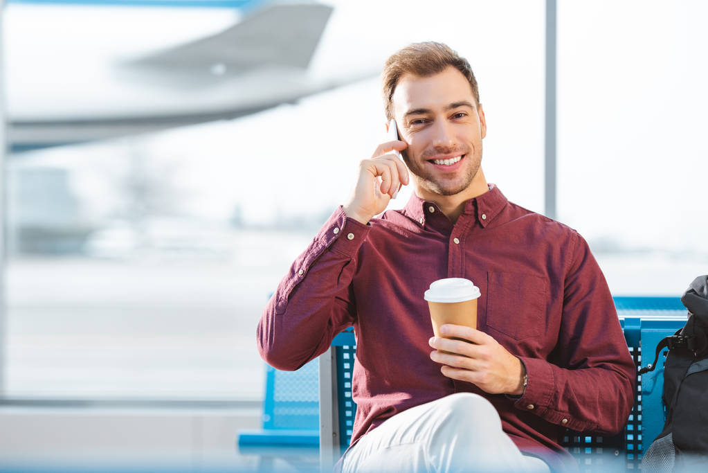 homem alegre falando no smartphone e segurando copo de papel na mão no aeroporto
  - Foto, Imagem
